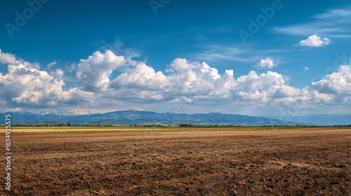 A plowed field under a blue sky with white clouds and mountains in the distance on a sunny day