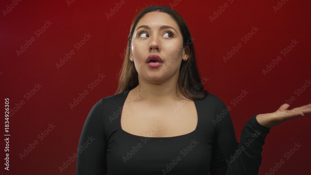 Fototapeta premium Young hispanic woman wearing a fitted black long sleeve top with palm raised and eyes closed against a red studio wall; indifference.