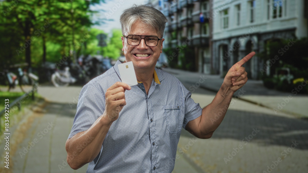 Fototapeta premium Man with grey hair holding a plain creditcard, smiling and pointing finger toward the right on a tree lined street; trust confidence promotion.