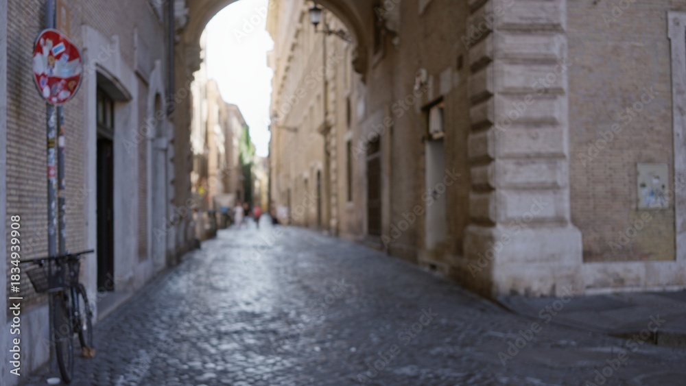 Fototapeta premium Narrow cobblestone street framed by historic brick buildings and archway, softly defocused street; background backdrop copyspace calm.