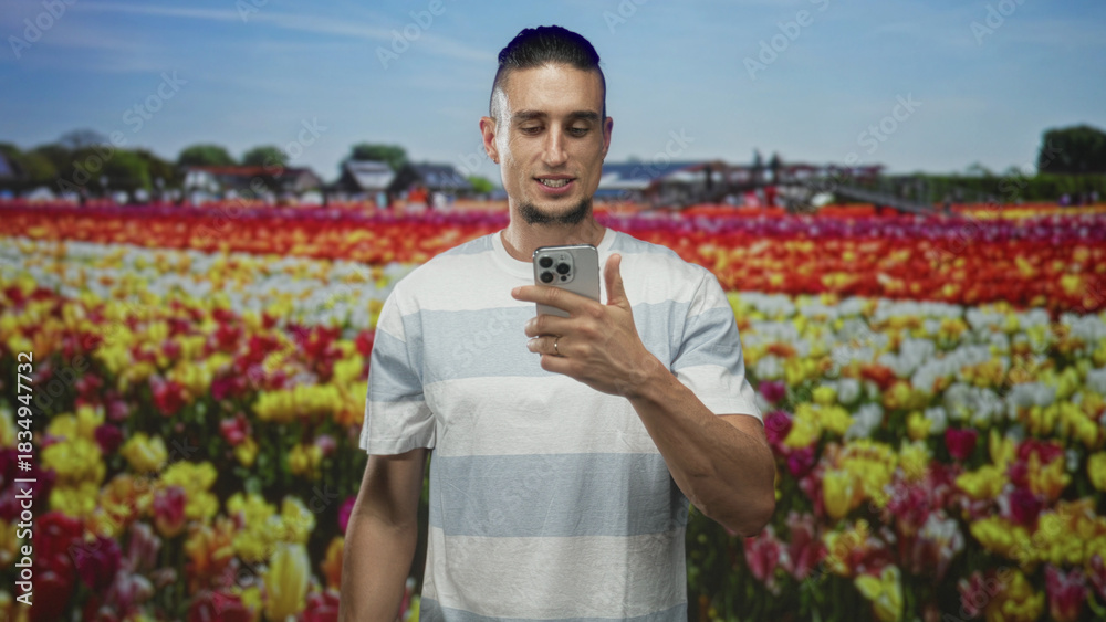 Fototapeta premium Man holding smartphone and looking down at the phone, hand visible over device against a studio tulip field backdrop; contemplation.