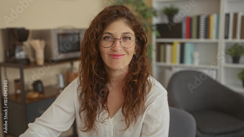 Foto Woman with round glasses smiles at camera in an office with bookshelf and chair, showing freckled face and relaxed posture; calm confidence