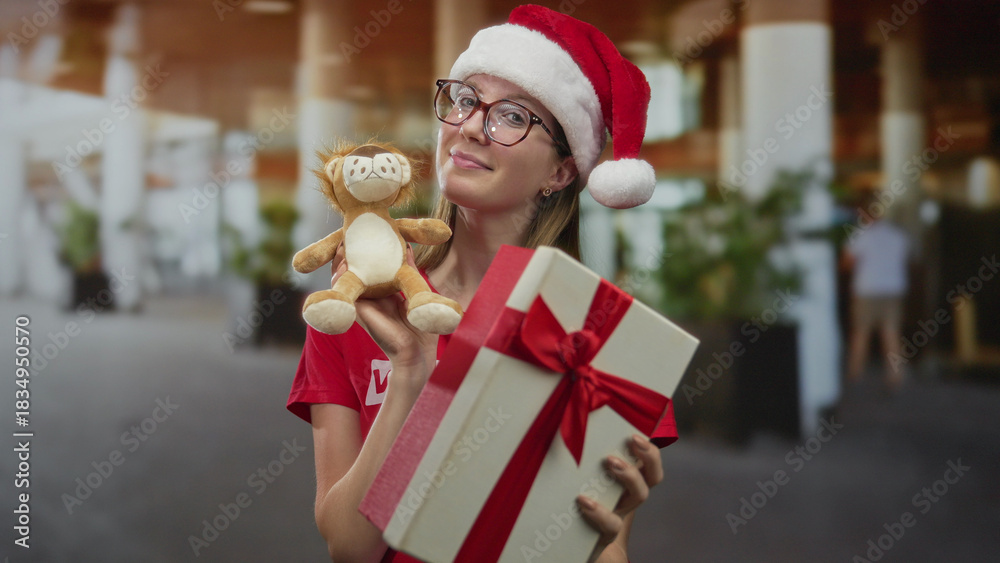 Fototapeta premium Woman wearing santa hat smiles and holds gift box and plush toy in building; holiday generosity giving.