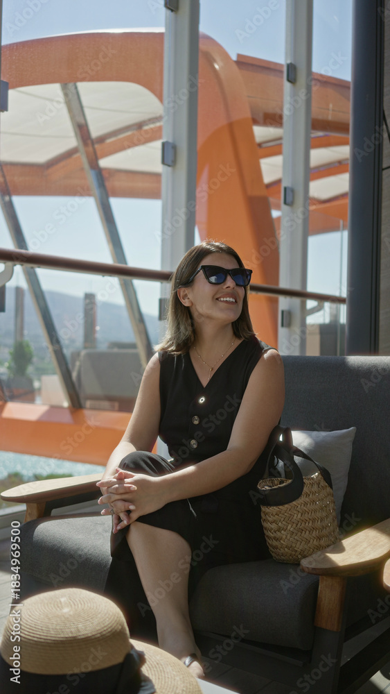 Fototapeta premium Woman holding sunglasses on cruise terrace lounge chair with straw bag and hat beside her while she sits and reaches toward hat; travel leisure calm.