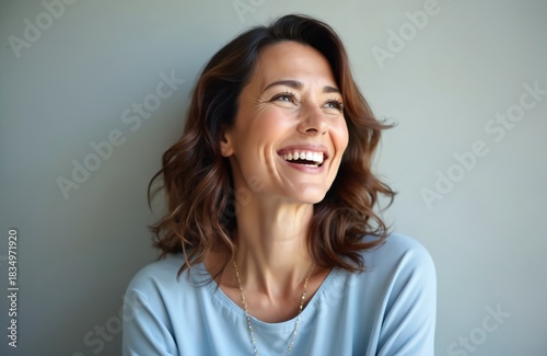 Happy middle-aged woman portrait smiling looking away. Attractive female with beautiful face expression, open mouth shows white teeth. Brunette lady wears blue shirt on grey studio background.