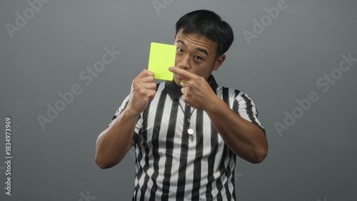 Young chinese referee holds yellow card and points finger while wearing striped shirt in grey studio; authority.