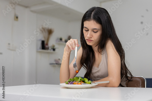 Asian women eating salad with unhappy expression, showing lack of appetite or emotional conflict related to healthy eating, dieting pressure or mental struggle around food choices at home.