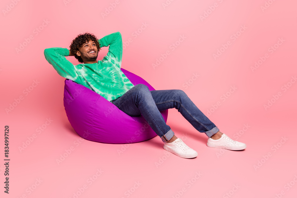 Fototapeta premium Young man relaxing on pink background sitting on a purple bean bag wearing green tie dye sweater jeans and white sneakers smiling