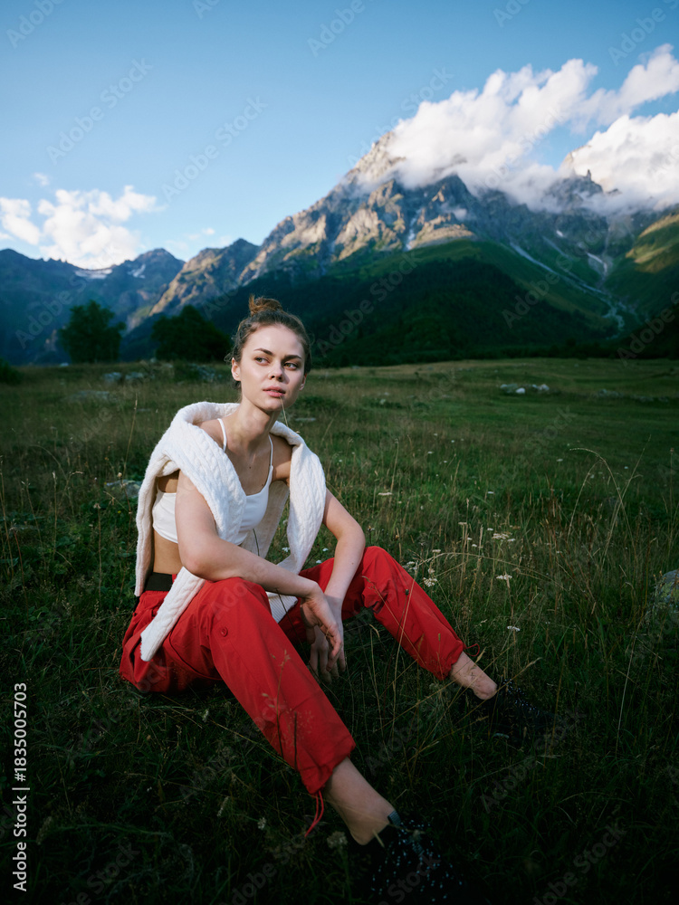 Fototapeta premium Woman sitting in a grassy meadow with towering mountains in the background, wearing a white top and red pants, calm mood, natural light and expansive alpine scenery for fashion and outdoor lifestyle