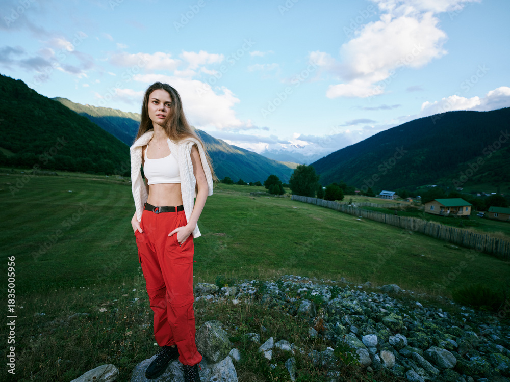 Fototapeta premium Woman outdoors on a grassy field with mountains in the background, wearing red pants and white top with a vest, casual fashion amid a scenic landscape and blue sky