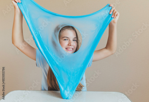 Playful girl peeking through a hole in a sheet of bright blue slime stretched in front of her face. Fun sensory toy