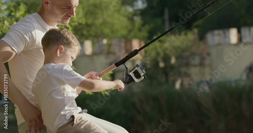 Father and son smile while sitting together on wooden pier holding fishing rod. Parent helps adorable child to pull small fish from sparkling surface of lake
