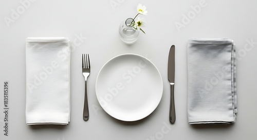 Elegant table setting for a formal meal, featuring a white plate, cutlery, and a delicate flower in a vase, all arranged on a plain grey background