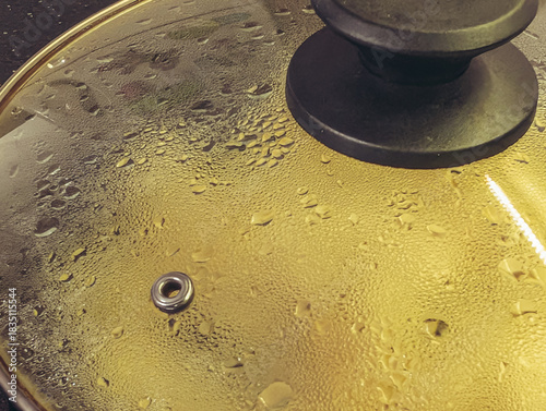 Close-up of steam and droplets forming on a glass saucepan lid with a yellow light reflection.