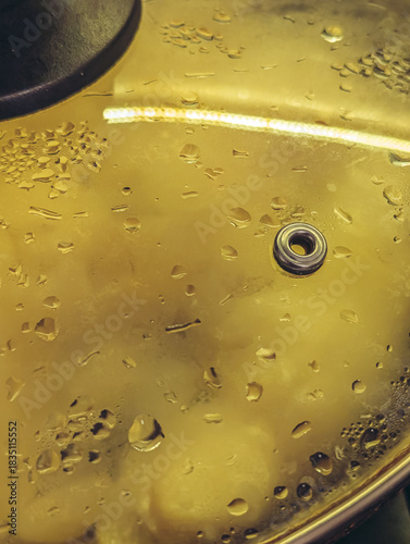 Close-up of steam and droplets forming on a glass saucepan lid with a yellow light reflection.