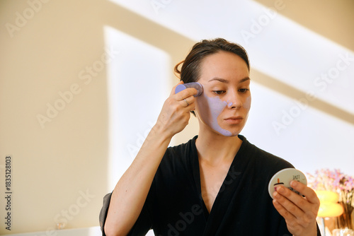 A young woman applies a clay mask in a stick to her face, looking in a mirror. Facial skin care at home