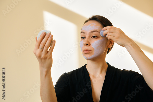 A young woman applies a clay mask in a stick to her face, looking in a mirror. Facial skin care at home