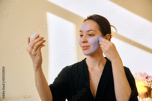 A young woman applies a clay mask in a stick to her face, looking in a mirror. Facial skin care at home