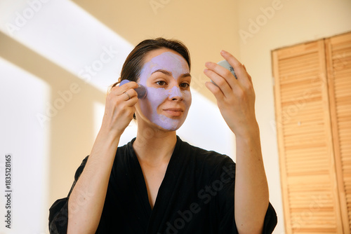 A young woman applies a clay mask in a stick to her face, looking in a mirror. Facial skin care at home