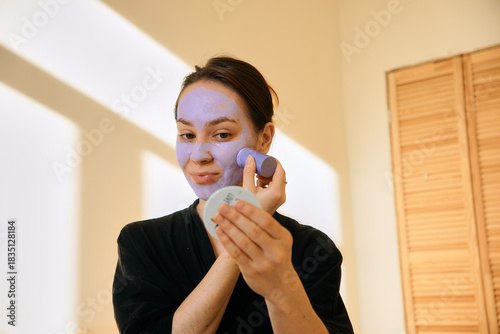 A young woman applies a clay mask in a stick to her face, looking in a mirror. Facial skin care at home