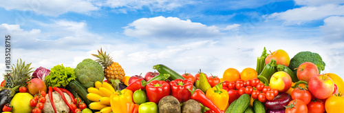 Assorted fresh fruits and vegetables arranged outdoors under blue sky with scattered clouds