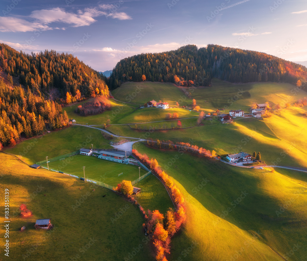 Fototapeta premium Aerial view of green alpine valley at sunset with scattered villages, fields, forests and rolling hills in Dolomites, Italy. Scenic rural mountain landscape with colorful autumn nature. Top drone view