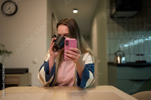 Woman drinking coffee and browsing smartphone at kitchen table