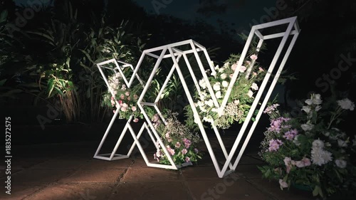 Wallpaper Mural Illuminated metal letters forming “XV” stand on garden path at night, with open geometric frames filled with pastel flowers, creating modern decorative backdrop for Sweet 15 quinceañera celebration Torontodigital.ca