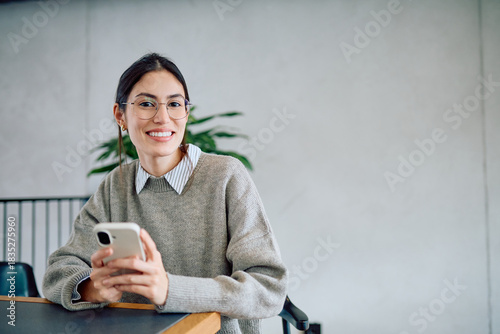 Photos Young woman wearing glasses and a sweater, smiling and holding a smartphone, loo