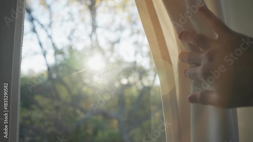 Sunlight shining behind window curtain as hand draws drape open as sun flares. Silhouette of male hand reveals sun behind window inside house or apartment overlooking nature background or garden