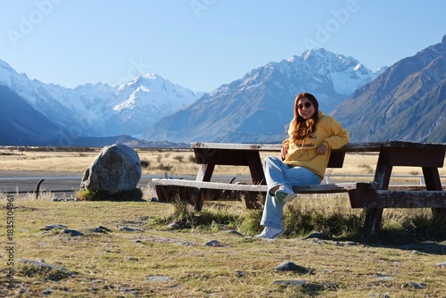 Mount Cook Airport, NZ - May 24, 2025: A person in a yellow hoodie smiles while seated on a wooden picnic bench. The vast, snow-capped peaks of the Southern Alps form a magnificent, clear-day backdrop