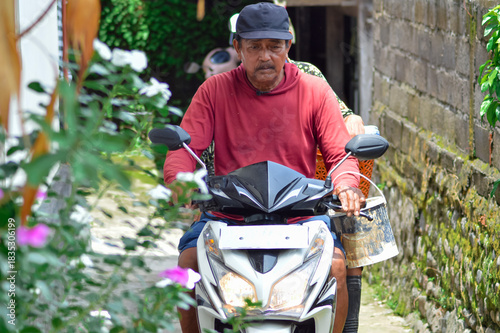 A front close-up view of a middle-aged villager couple riding their motorbike into a narrow residential path, the father drives forward while the mother rides pillion carrying a bucket.