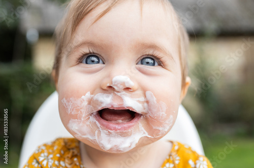 portrait of face, happy little girl under one year old, eating yogurt on her own and getting dirty. child's face is smeared with food. Happy, sincere joy, smile. A cheerful childhood. Daily routine.
