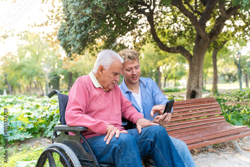 Young adult grandson showing smartphone to senior man in wheelchair while sitting together in park