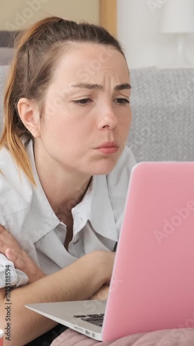 Bored young woman resting chin on hands while looking at laptop screen, getting annoyed and disappointed while lying on floor at home. Vertical video.