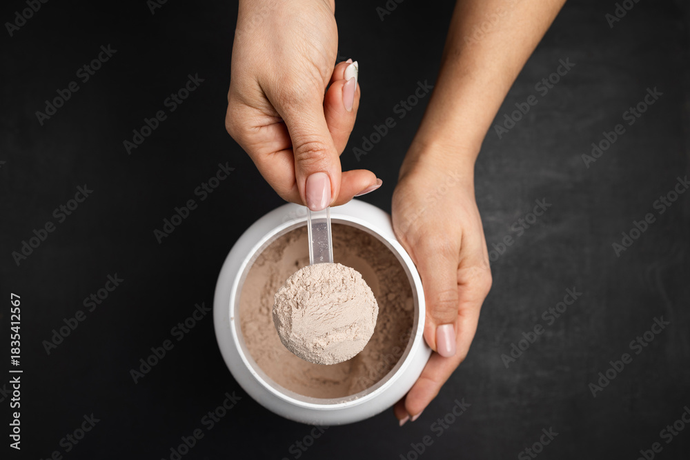 Fototapeta premium Top view of female hand holding measuring scoop with chocolate whey protein powder above plastic jar on dark background, concept of sports nutrition and dietary supplements