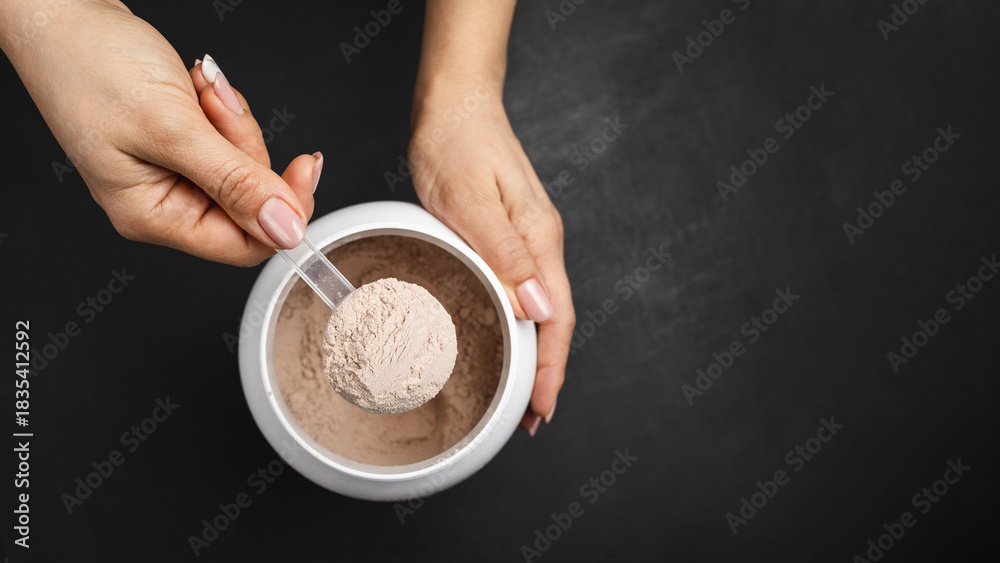 Fototapeta premium Top view of female hand holding measuring scoop with chocolate whey protein powder above plastic jar on dark background with copy space, concept of sports nutrition and dietary supplements