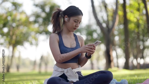 Young Asian adult woman connects with smartphone and wireless earbuds in park setting healthy lifestyle wellness tech outdoor fitness activity