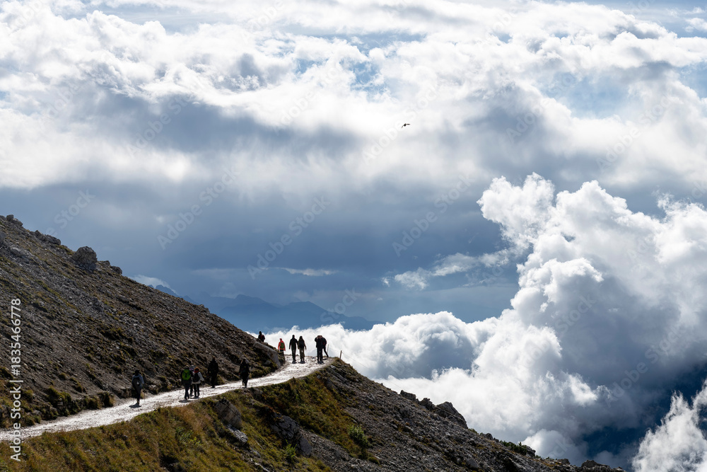 Fototapeta premium Hikers on sun glistening hiking path towards Tre Cime di Lavaredo peaks (Drei Zinnen) with cloudscape of dark storm clouds around peaks and white sun lit clouds in valley in Dolomite Alps, Italy