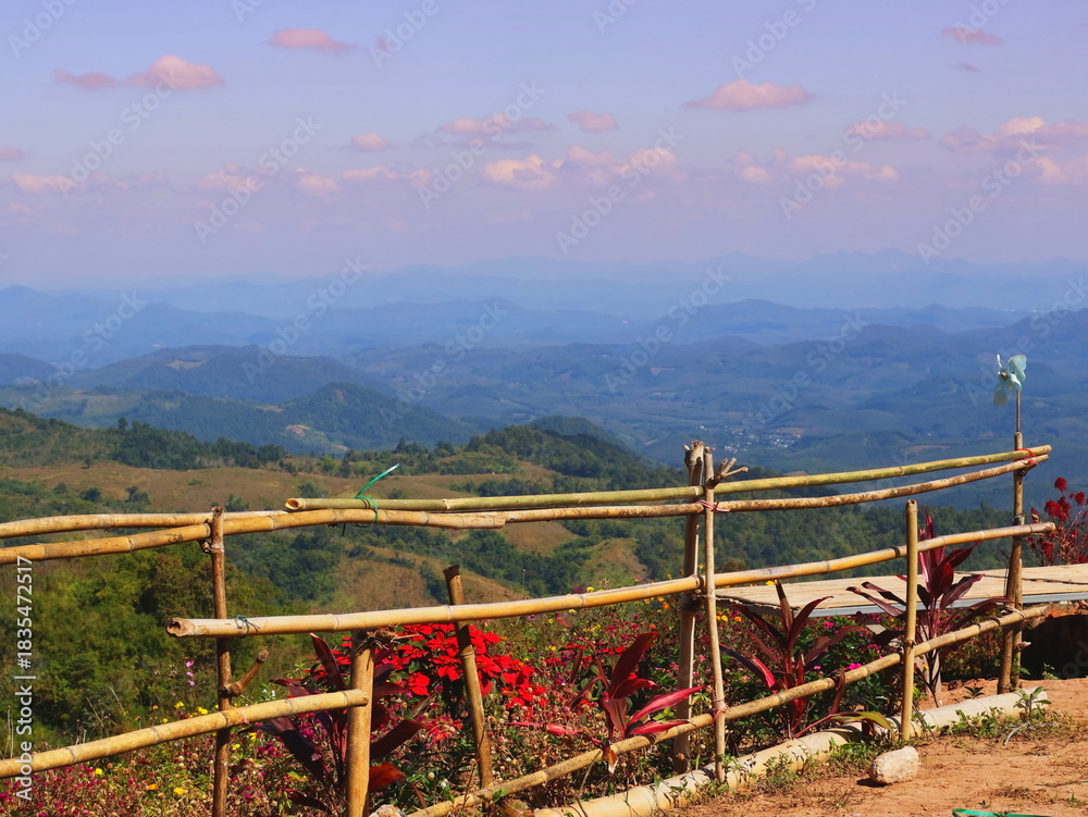 Fototapeta premium The landscape features a bamboo fence and bright red flowers, with layered mountains shrouded in a light mist in the background.