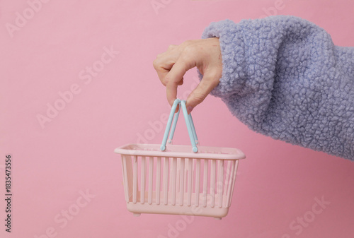 Female hand holding a toy supermarket shopping basket on a pink background