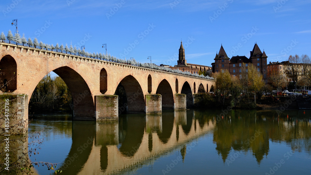 Fototapeta premium Scenic View of Historic Stone Bridge Spanning Over Serene River with Cityscape Background Under Clear Blue Sky in Montauban Setting