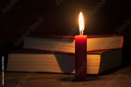 A red candle burns on a wooden table, its warm glow illuminating two books behind it. A soft, calm atmosphere, ideal for themes like reading, coziness, and winter evenings.