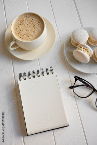 White wooden table with warm coffee, colorful macarons, open spiral notebook and reading glasses, cozy creative planning scene for work, study or a relaxing morning break
