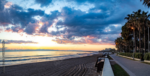 Fototapeta Naklejka Na Ścianę i Meble -  A vast, empty beach with perfectly raked sand under a dramatic orange sunset sky, ready for the new season. A serene and tranquil panorama of the Mediterranean coast. Side, Antalya, Turkey.

