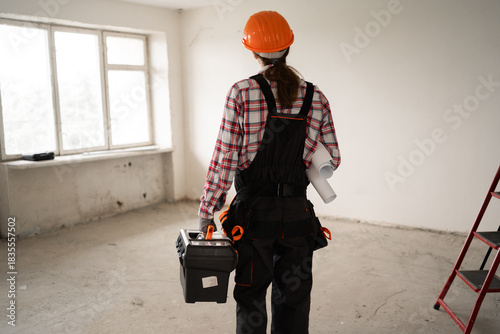 Rear view of male repair worker or engineer in hard hat and tool belt holding toolbox preparing to work in new modern house