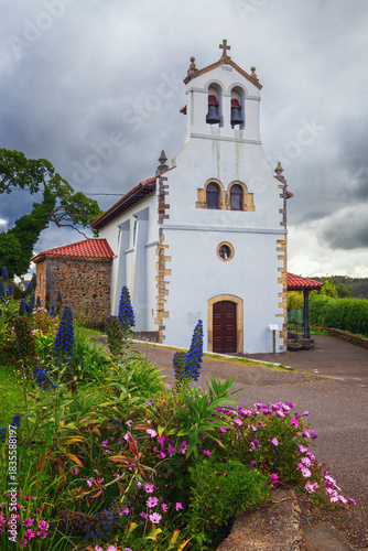 Santiago del Monte parish church, way of st. james, with a white facade and a bell tower with two bells, surrounded by colorful flowers and vegetation, Asturias, spain.