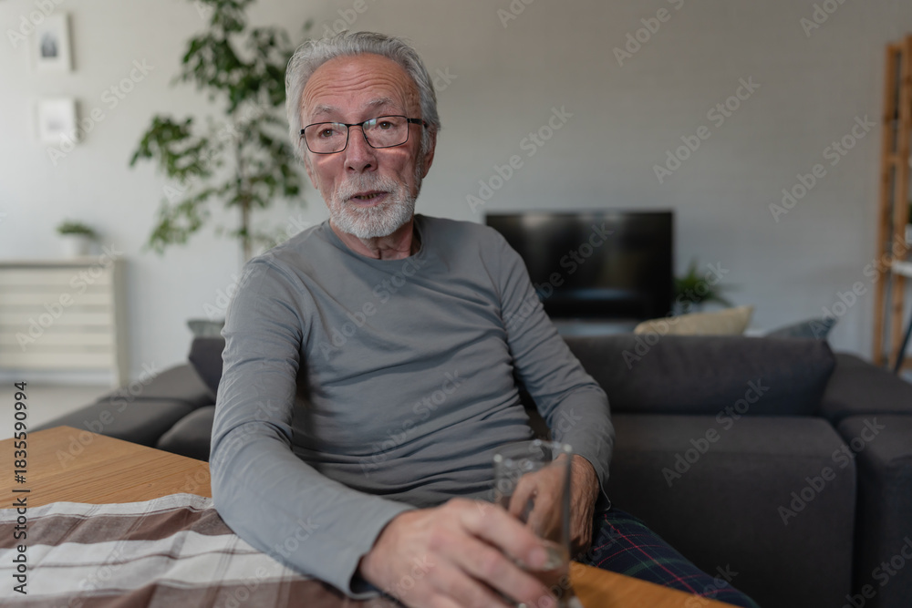 Fototapeta premium Elderly man sitting at a table in a cozy living room, holding a glass of water and looking relaxed