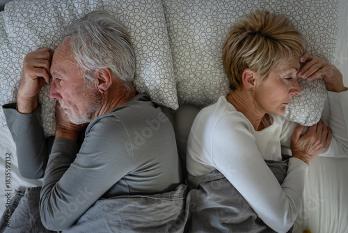 Senior couple lying back-to-back in bed, appearing distant and thoughtful, covered with gray blankets in a quiet bedroom