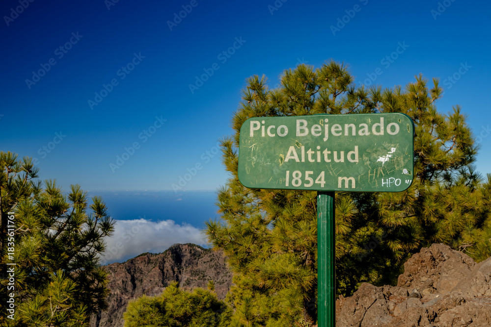 Obraz premium Señal verde en el Pico Bejenado, La Palma, indicando 1854 m de altitud. Se ve la cresta rocosa y un mar de nubes bajo el cielo azul claro.